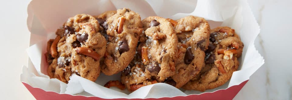 Top-down shot of four cookies in a red basket. Cookies topped with salt, pretzels, chocolate chips.  