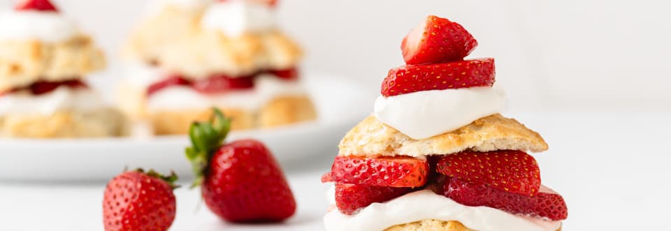 Side profile of biscuit with strawberries and whipped cream and two strawberries on the left side upon a white countertop.