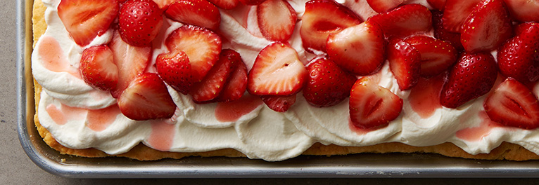 A sheet pan dessert topped with a thick layer of whipped cream and a generous amount of fresh, sliced strawberries drizzled with strawberry syrup.