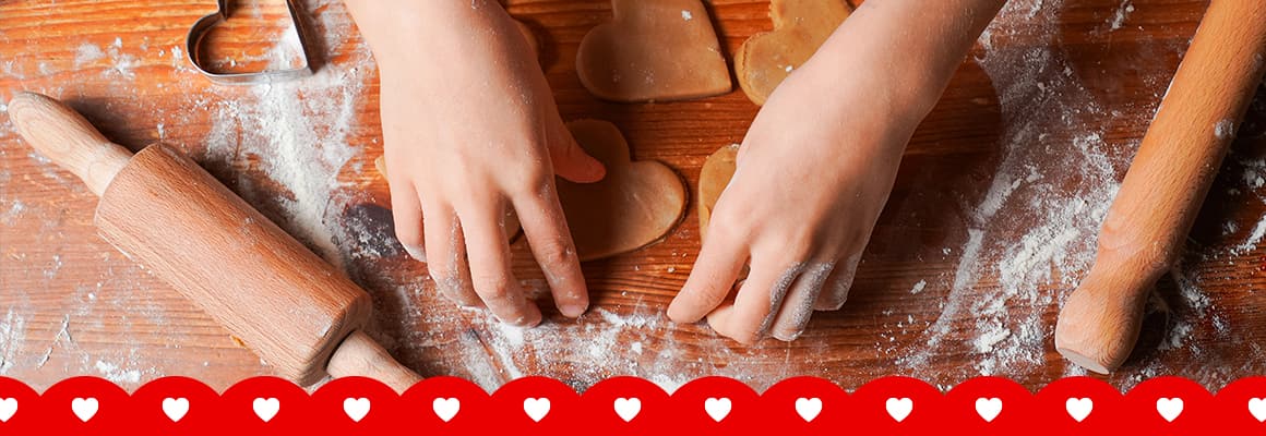 Two hands over, a heart shaped cookie dough, rolling pin, heart shaped cookie cutters, on a table dusted with flour