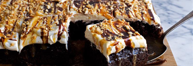 Overhead shot of chocolate sheet cake with whipped cream, chocolate, caramel drizzle, and crushed peanuts served on a cutting board.