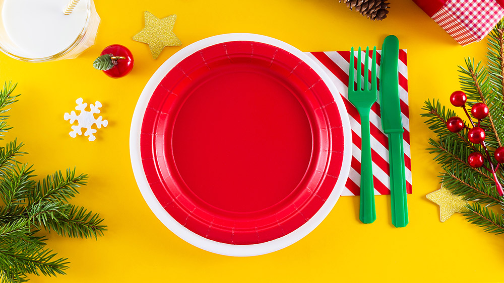 Place setting with a yellow table cloth, red plate, green fork and knife, red and white striped napkin and pine boughs as decorations