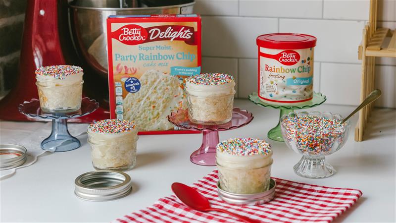 Mini sprinkle-topped cakes in jars with cake mix and frosting on a kitchen counter.