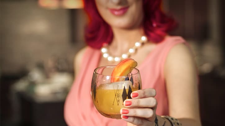 woman with red hair, vintage pink dress and wrist tattoo holding an orange cocktail with orange slice in a retro lowball glass