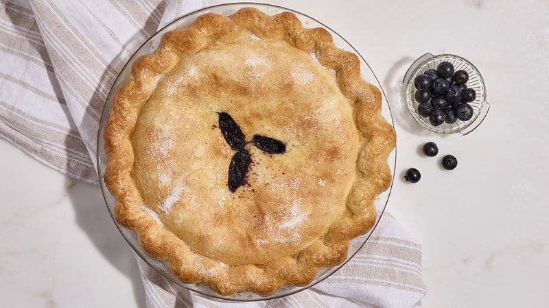 A golden‑brown blueberry pie with a sugared crust and three slits in the center, sitting on a striped towel with a small bowl of fresh blueberries beside it.